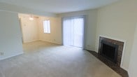 This single-floor space offers a clean, functional layout designed for comfortable everyday living. The main living area is generously sized, featuring plush wall-to-wall carpeting that adds warmth underfoot. A standout feature of the room is the brick-surround fireplace, which serves as a classic focal point and brings a cozy, traditional character to the space. Natural light fills the room through a large sliding glass door dressed with vertical blinds, which opens to an outdoor area and provides a bright, airy atmosphere. An additional window on the adjacent wall further enhances the natural light throughout the day. A ceiling fan with a light fixture is mounted overhead, offering both comfort and illumination. The walls are finished in a neutral, light tone that complements the soft carpeting and keeps the overall aesthetic clean and versatile. The space is well-suited for a variety of furniture arrangements, allowing for a personalized and practical setup. The combination of natural light, classic fireplace, and open layout gives this space a welcoming and livable ambiance throughout.