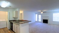 This single-floor space showcases an open-concept design that seamlessly connects the kitchen and living area, creating a cohesive and functional layout. The kitchen features white cabinetry, a dark granite-style countertop, and a peninsula that doubles as a breakfast bar, providing both additional prep space and casual seating. A double-basin sink is integrated into the peninsula, adding practicality to the layout.

The adjoining living area is finished with neutral-toned carpeting that adds warmth underfoot. A ceiling fan with a light fixture is centrally positioned, ensuring comfortable airflow and ambient lighting throughout the space. A tile-surround fireplace serves as a notable focal point along one wall, offering both visual appeal and a cozy gathering spot.

Natural light fills the living area through a set of glass patio doors, which also provide direct access to an outdoor area, enhancing the sense of openness. The walls throughout are finished in a clean, neutral tone, creating a bright and versatile backdrop. A hallway visible from the kitchen suggests additional rooms beyond the main living space.