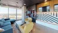 This striking high-rise amenity space offers an open, single-floor layout designed for social gathering and entertainment, elevated by sweeping panoramic city views visible through expansive floor-to-ceiling windows. The polished concrete flooring and exposed concrete ceiling provide an industrial foundation that is complemented by warm wood slat wall paneling, creating a dynamic contrast of textures throughout the space.

The main open area is anchored by a full-size pool table with a bold yellow felt surface, positioned to take full advantage of the city skyline backdrop. Adjacent to it, a bar-height counter with stools provides an additional social hub framed by a vibrant pink accent structure. A dedicated lounge zone features a large sectional sofa and coffee table, bathed in natural light from the surrounding glass walls, offering unobstructed elevated views of the cityscape.

A secondary seating area is distinguished by its decorative wall of recessed display niches showcasing colorful sculptural accents, a mounted flat-screen television, and a striking geometric mural. Mid-century-inspired accent chairs and a low coffee table complete this intimate corner. The entry area features a combination of wood paneling and dark painted surfaces, with an accessible lift and staircase providing level changes within the space. Recessed lighting throughout ensures a well-lit, welcoming atmosphere at all hours.