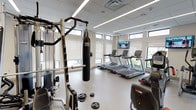 This fitness center offers a comprehensive workout environment designed to accommodate a wide range of exercise disciplines. The space is housed within a clean, modern interior featuring a drop tile ceiling with linear LED lighting that evenly illuminates the entire floor. A row of clerestory windows runs along the upper perimeter of the walls, allowing natural light to filter in while maintaining privacy, and providing glimpses of greenery outside.

The cardio zone is well-appointed with multiple treadmills, elliptical trainers, and stationary bikes, all positioned to face wall-mounted flat-screen televisions for entertainment during workouts. The strength training area includes a multi-station cable machine by Hoist, a dedicated free weights section with weight plates, and a heavy PRO punching bag suspended from a power rack — catering to both traditional and combat-style training.

Functional training is further supported by a rack of stability and medicine balls in assorted sizes, resistance accessories, and additional cable attachment stations. Full-length mirrors line one wall, enhancing the sense of openness and allowing users to monitor their form. The light-toned flooring and neutral white walls contribute to a clean, airy atmosphere throughout. The facility is thoughtfully laid out to balance equipment density with ample open space for movement and stretching.