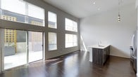 This single-floor space showcases a striking contemporary design characterized by dramatically high ceilings that amplify the sense of openness throughout. The main living area is flooded with natural light, courtesy of an expansive wall of floor-to-ceiling windows and a glass sliding door that opens directly to an outdoor patio or terrace, blurring the boundary between interior and exterior living.

The flooring throughout features rich, dark hardwood that adds warmth and contrast against the crisp white walls and bright ceiling. Recessed lighting complements the abundant natural light, ensuring the space remains well-lit at all hours.

Seamlessly integrated into the open-concept layout, the kitchen is positioned along one wall and features sleek, dark wood cabinetry with a clean white countertop and an undermount sink with a modern faucet. A stainless steel dishwasher is tucked neatly beneath the counter. The minimalist design of the kitchen complements the overall aesthetic of the space.

The result is a bright, open, and thoughtfully designed interior that balances modern finishes with functional living.
