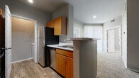 This single-floor residence offers a practical and comfortable layout designed to maximize functionality and livability. The kitchen is a standout feature, equipped with warm-toned wood cabinetry, white countertops, and a full suite of stainless steel and black appliances including a refrigerator, dishwasher, microwave, and range. Wood-look flooring runs throughout the kitchen, and a peninsula with a granite-style countertop provides a convenient transition into the adjacent living area. The living space is carpeted and benefits from abundant natural light streaming through large windows fitted with vertical blinds, creating a bright and open atmosphere. The spacious bedroom continues the carpeted flooring and includes a ceiling fan, multiple windows, and a walk-in closet with built-in shelving, offering generous storage. Two full bathrooms mirror each other in design, each featuring a tub-shower combination with tiled surround, a single-sink vanity with wood cabinetry and granite-style countertop, a large mirror, and wood-look flooring. A separate laundry area with washer and dryer hookups adds further convenience. The neutral gray wall palette throughout gives the space a clean, modern feel that is move-in ready.