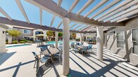 Located at 9353 W Twain Ave in Las Vegas, Nevada, this outdoor amenity area showcases a resort-style courtyard designed for relaxation and leisure under the Nevada sun. The centerpiece is a generously sized freeform swimming pool with crystal-clear blue water, surrounded by an expansive pool deck lined with lounge chairs for sunbathing. Adjacent to the pool, a dedicated spa provides a more intimate retreat, set amid mature trees and manicured landscaping that add welcome shade and a lush, inviting atmosphere.

A standout architectural feature is the substantial pergola structure, supported by sturdy stucco-finished columns and fitted with an open-beam overhead lattice that casts striking geometric shadows across the concrete patio. Beneath the pergola, a variety of seating arrangements — including ornate metal bistro tables and cushioned chairs — create comfortable spaces for outdoor dining and socializing. Additional umbrella-shaded seating clusters are scattered throughout the courtyard, offering flexible options for gathering.

The surrounding buildings reflect a classic Spanish Revival architectural style, characterized by red clay tile roofs, arched entryways, stucco exteriors, and wrought-iron balcony railings. Wall-mounted lantern-style light fixtures accent the exterior facades. The entire outdoor space is enclosed by decorative iron fencing, providing a sense of privacy and security while maintaining an open, airy feel throughout the community courtyard.