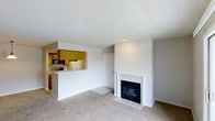 This single-floor residence offers a practical and comfortable layout designed for easy living. The living room serves as the heart of the space, featuring wall-to-wall carpeting that adds warmth underfoot and a classic white-mantled fireplace framed in tile, creating a welcoming focal point. A textured ceiling runs throughout, adding subtle character to the interior.

The kitchen is thoughtfully connected to the living area via a pass-through opening, promoting an open and social atmosphere. Warm wood cabinetry lines the kitchen walls, complemented by essential appliances including a refrigerator, microwave, and range. A tiled countertop bar along the pass-through provides a convenient casual dining or prep surface, while the kitchen floor transitions to tile, offering easy maintenance in the cooking area.

A doorway visible from the main living space leads further into the residence, hinting at additional private areas beyond. The overall color palette is neutral and clean, with white walls and light tones throughout that enhance the sense of brightness and openness. This well-organized single-level layout balances functionality with comfort, making it an inviting and efficient living space.