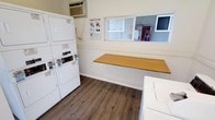 This single-floor laundry facility is designed for practical, efficient use. The room features stacked washer and dryer units along one wall, maximizing the available floor space while accommodating multiple loads simultaneously. A standalone washer is also present, offering additional capacity. A wide, wood-toned folding counter runs along the adjacent wall, providing a dedicated surface for sorting and folding laundry. The flooring consists of durable wood-look planks that are easy to maintain. A wall-mounted air conditioning unit ensures comfort during use. Natural light filters in through a horizontal window, and a pass-through window on the same wall suggests a connection to an adjacent service or office area. Instructional signage is posted on the wall, offering guidance on proper drying techniques. The space is finished with light-colored walls that contribute to a clean, utilitarian atmosphere. Overall, the layout is straightforward and functional, prioritizing ease of use and efficient workflow for laundry tasks.