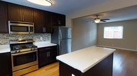 This two-floor space offers a thoughtfully designed layout that balances functionality and style. The kitchen is a standout feature, showcasing rich dark cabinetry with sleek hardware, bright white countertops, and a full suite of stainless steel appliances, including a gas range, built-in microwave, and refrigerator. A decorative mosaic tile backsplash adds visual interest and texture to the cooking area, while warm hardwood flooring grounds the space with a natural, inviting feel. A central kitchen island with a clean white surface provides additional prep space and serves as a natural gathering point. The kitchen flows seamlessly into an adjacent living area, which is illuminated by a ceiling fan with an integrated light fixture and benefits from natural light through a nearby window. The neutral wall tones throughout create a calm, versatile backdrop suitable for a variety of décor styles. The space spans two floors, offering distinct zones for living, entertaining, and private retreat, making it a practical and comfortable environment for everyday living.
