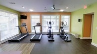 This fitness center offers a clean, functional environment designed for effective workouts. The space is outfitted with a selection of cardio equipment, including multiple treadmills, an elliptical cross-trainer, and a stationary bike, all arranged to allow comfortable use and easy movement between machines. Durable, textured rubber flooring covers the workout area, providing secure footing during exercise, while a section of smooth hardwood-style flooring is visible near the entry area. Large windows along the main wall flood the room with natural light and offer pleasant views of the surrounding greenery, creating an open and energizing atmosphere. Warm orange and soft green accent walls add a lively, motivating character to the space. A ceiling fan ensures comfortable air circulation during workouts, complementing the recessed lighting overhead. A wall-mounted flat-screen television is positioned for easy viewing from the cardio equipment. The space is thoughtfully laid out to maximize usability while maintaining an uncluttered, welcoming feel suited for daily fitness routines.