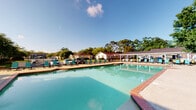 This outdoor pool facility in Mobile, Alabama, offers a well-appointed aquatic retreat designed for both relaxation and recreation. The centerpiece is a large, rectangular swimming pool with clear turquoise water, framed by a broad concrete and brick-accented deck that provides ample space for sunbathing and socializing. Rows of teal lounge chairs are arranged along the pool's perimeter, offering comfortable seating for residents throughout the day.

The area is enclosed by ornamental black iron fencing, which provides both security and a classic aesthetic. A covered pavilion runs along one side of the pool, supported by white columns and equipped with pendant lantern-style light fixtures, offering a shaded gathering space with direct views of the pool. The pavilion's ceiling is finished in white, contributing to a clean, polished look.

Mature, lush trees surround the perimeter of the space, providing natural shade and a sense of seclusion. The adjacent single-story building features light-colored vinyl siding and standard windows, housing supporting amenities. Safety equipment, including life rings, is visibly positioned along the fencing. Under open Alabama skies, this pool area delivers a functional and inviting outdoor environment suited for warm-weather enjoyment.