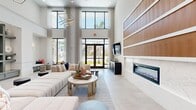 This elegantly designed single-floor amenity space offers a generous, open layout that seamlessly blends comfort and sophistication. The defining architectural feature is the dramatic double-height ceiling, which amplifies the sense of openness and is complemented by floor-to-ceiling windows and glass entry doors that flood the interior with natural light and frame views of lush outdoor greenery.

The lounge area serves as the centerpiece, furnished with plush, tufted sectional sofas accented by vibrant throw pillows. Statement geometric pendant light fixtures hang overhead, adding a sculptural, artistic element. A built-in wall of open shelving provides both display space and visual structure, while a sleek linear fireplace set into a warm wood-paneled accent wall adds a refined focal point.

Adjacent to the lounge, a dedicated game room features a full-sized pool table and a shuffleboard table, with additional seating positioned around the perimeter for spectators. Abstract artwork adorns the crisp white walls throughout.

A stylish kitchenette area offers sage-green cabinetry, a quartz waterfall island, a textured tile backsplash, and stainless steel appliances. Woven rattan pendant lights complete the coastal-modern aesthetic. Light-toned large-format tile flooring unifies the entire space.