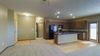 This single-floor space is designed with an open-concept layout that connects the kitchen and living areas in a cohesive, functional flow. The kitchen is a standout feature, showcasing rich dark wood cabinetry that extends to the ceiling for ample storage, complemented by dark countertops and a full suite of black appliances, including a refrigerator with an ice and water dispenser, a range, and an overhead microwave. A peninsula-style kitchen island provides additional workspace and serves as a natural dividing point between the kitchen and the adjacent living area, and features a built-in sink for added convenience. Recessed lighting throughout the kitchen ensures the space is well-lit, while a ceiling fan in the living area promotes air circulation. The living area transitions from hardwood-style flooring in the kitchen to plush carpet, adding warmth and comfort underfoot. Neutral-toned walls throughout give the space a clean, welcoming ambiance. A hallway visible from the main living area leads to additional rooms, hinting at further private spaces beyond. The overall design balances practicality with a cohesive aesthetic, making it a comfortable and well-appointed living environment.
