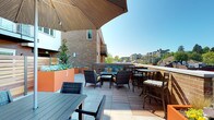 This rooftop terrace offers a well-appointed outdoor amenity space designed for both relaxation and social gatherings. The terrace features a striking wood-paneled curved shade canopy that serves as a defining architectural element, providing shelter while adding warmth and visual interest to the space. The layout is thoughtfully organized into multiple distinct seating zones. A central lounge area is furnished with wicker chairs and a loveseat accented with colorful teal and yellow cushions, arranged around a low coffee table — ideal for casual conversation. Nearby, bar-height tables with stools offer an alternative seating option for smaller groups. Additional dining-style tables expand the space's capacity for entertaining. Vivid orange raised planters introduce greenery and a bold pop of color throughout the terrace, enhancing the lively, welcoming atmosphere. The flooring combines large-format tile with wood-toned deck sections, adding textural contrast underfoot. The terrace enjoys ample natural light under open skies and offers panoramic views of the surrounding urban skyline and neighboring buildings, creating an inviting connection to the broader cityscape. Overall, the space balances modern design with functional comfort.