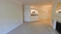 This single-floor residence is designed with an open-concept layout that promotes a sense of spaciousness and ease of movement. The main living area is finished with plush, light-toned wall-to-wall carpeting that adds warmth and comfort underfoot. Neutral cream-colored walls throughout contribute to a bright, clean aesthetic that suits a variety of furnishing styles.

The kitchen is positioned centrally and features a pass-through counter with a dark contrasting countertop, creating a natural divide between the cooking and living spaces while maintaining visual openness. White cabinetry lines the kitchen walls, offering ample storage, and is complemented by stainless steel appliances, including a microwave and range. Recessed overhead lighting illuminates the kitchen workspace effectively.

Adjacent to the living area, a pendant light fixture hints at a dedicated dining space, adding a subtle decorative touch. Crown molding details along the upper walls lend a refined, finished look to the interior. The overall ambiance is clean, neutral, and move-in ready, offering a versatile canvas for personalization.