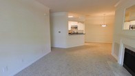 This single-floor residence is designed with an open-concept layout that promotes a sense of spaciousness and ease of movement. The main living area is finished with plush, light-toned wall-to-wall carpeting that adds warmth and comfort underfoot. Neutral cream-colored walls throughout contribute to a bright, clean aesthetic that suits a variety of furnishing styles.

The kitchen is positioned centrally and features a pass-through counter with a dark contrasting countertop, creating a natural divide between the cooking and living spaces while maintaining visual openness. White cabinetry lines the kitchen walls, offering ample storage, and is complemented by stainless steel appliances, including a microwave and range. Recessed overhead lighting illuminates the kitchen workspace effectively.

Adjacent to the living area, a pendant light fixture hints at a dedicated dining space, adding a subtle decorative touch. Crown molding details along the upper walls lend a refined, finished look to the interior. The overall ambiance is clean, neutral, and move-in ready, offering a versatile canvas for personalization.