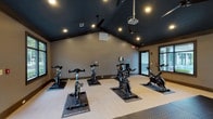 Located at 6721 Washington Ave in Ocean Springs, Mississippi, this single-floor fitness studio is thoughtfully designed to support a wide range of workout routines. The space is defined by its striking vaulted ceilings finished in a deep navy/charcoal tone, complemented by recessed lighting and ceiling fans that maintain airflow and comfort throughout sessions.

The studio is divided into two functional zones. The cycling area accommodates multiple Bodycraft stationary spin bikes arranged on protective rubber mats, with a projector mounted overhead for guided class experiences. A water station is conveniently positioned nearby. Glass doors open directly to an outdoor area, offering natural light and views of the surrounding pool and grounds.

The strength and functional training zone features dual dumbbell racks stocked with a comprehensive range of free weights, a large fixed projection screen mounted against a wood-paneled accent wall, medicine balls, stability balls, step platforms, and interlocking rubber floor tiles for durability and safety. Large windows throughout the studio allow ample natural light to fill the space while providing views of the exterior.

The overall design balances a modern, high-performance aesthetic with practical functionality, making it a versatile and well-appointed fitness facility suitable for both individual training and group exercise.