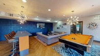 This single-floor community lounge space is designed with a bold, contemporary aesthetic that combines deep navy blue and vivid orange accents throughout. The open-plan layout seamlessly connects a social lounge area, a recreation zone, and a fully equipped kitchen, creating a cohesive and energetic environment ideal for gathering and entertainment.

The lounge area serves as the centerpiece of the space, featuring a large tufted sectional sofa arranged around a geometric-patterned area rug and a round coffee table. Abstract artwork adorns the walls, complemented by eye-catching sputnik-style chandeliers that cast a warm, ambient glow. A striking navy blue textured accent wall with a raised geometric panel design anchors one end of the room, adding architectural depth and visual interest.

Adjacent to the lounge, a billiards table with a bright orange felt surface sits beneath its own sputnik chandelier on a herringbone-patterned hardwood floor. A cozy seating nook with retro-style orange accent chairs is positioned near large windows that allow natural light to flood the space and offer views of the surrounding exterior.

The kitchen area features stainless steel appliances, white cabinetry with a mosaic tile backsplash, and an island bar with granite countertops and bar stool seating. Recessed lighting throughout ensures the entire space is well-lit, while the cohesive design elements tie the social, recreational, and culinary zones together into one dynamic, inviting space.