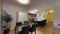 This single-floor space offers a thoughtfully designed open-concept layout that seamlessly connects the main living areas. The kitchen is well-appointed with dark espresso cabinetry, stainless steel appliances including a refrigerator, microwave, and range, and a light-toned countertop. A large kitchen island with an extended breakfast bar provides additional seating with stylish bar stools, making it an ideal spot for casual dining or entertaining. Pendant lighting hangs above the island, complementing the warm, ambient ceiling fixtures throughout the space. Adjacent to the kitchen, the dining area accommodates a full dining table with seating for multiple guests, finished with matching dark wood and black upholstered chairs. The warm-toned hardwood flooring flows continuously throughout, adding a cohesive and inviting feel to the space. A notable feature is the dedicated in-unit laundry room, visible through an open doorway, which houses a full-sized washer and dryer with shelving above. The overall ambiance is warm and functional, with neutral wall tones and a layout that maximizes the sense of openness and ease of movement throughout.