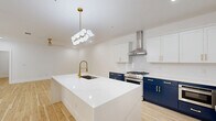 This thoughtfully designed 2-bedroom, 2-bathroom residence offers a cohesive, contemporary aesthetic carried throughout every room. The kitchen serves as the centerpiece of the open-concept living area, showcasing a striking two-tone design with crisp white upper cabinets and bold navy lower cabinetry, all accented with brushed gold hardware. A large waterfall quartz island with an undermount sink and a gold industrial-style faucet anchors the space, while a professional-grade stainless steel gas range with a wall-mounted hood, a French door refrigerator, and a modern globe chandelier complete the refined look. Light-toned hardwood flooring flows seamlessly from the kitchen and living area into the bedrooms, creating a bright, airy atmosphere throughout. The living area features floor-to-ceiling French doors that flood the space with natural light. Both bedrooms are generously proportioned, with clean white walls, recessed lighting, and ample natural light. The primary bedroom offers substantial square footage with dedicated closet space. The bathrooms are finished to a high standard, featuring marble-look tile walls and floors, frameless glass shower enclosures, navy vanity cabinetry with white quartz countertops, and backlit LED mirrors with gold fixtures — delivering a spa-like experience that mirrors the home's polished, modern character.