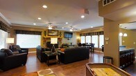 This single-floor space offers a warm and inviting atmosphere defined by rich hardwood flooring, neutral earth-toned walls, and a thoughtfully designed open layout. The living area serves as the centerpiece of the space, anchored by a striking stone fireplace with a warm wood mantle and framed artwork above. A wall-mounted flat-screen television complements the fireplace, making the area ideal for both relaxation and entertainment. Comfortable dark leather seating is arranged around a glass-top coffee table, creating a welcoming gathering space. Dual ceiling fans with integrated lighting are positioned throughout the main area, ensuring consistent airflow and comfort. Recessed lighting adds to the well-lit ambiance, supplemented by natural light streaming through multiple windows dressed with patterned valances and white blinds. The open-concept design flows seamlessly into a dining area, which features a dark wood table and chairs positioned near a large bay window that floods the space with daylight. A shuffleboard table visible at the edge of the room hints at additional recreational features. Crown molding throughout adds a refined architectural detail, tying the entire space together with a polished, cohesive finish.