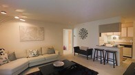 This single-floor space presents a thoughtfully designed open-concept layout that seamlessly connects the living area to the kitchen. The living room is anchored by a plush, light-toned sofa adorned with decorative cushions, complemented by a sleek dark coffee table resting atop a rich, dark area rug. A statement sunburst mirror serves as a striking focal point on the clean white walls, adding a touch of elegance alongside coordinating wall art. A compact accent bench provides additional seating near the hallway entrance, enhancing the room's functionality. The kitchen area features a raised bar-height counter with dark barstools, creating a casual dining and entertaining space. Neutral-toned cabinetry and appliances are visible beyond the counter, suggesting a well-equipped culinary space. Light-toned carpeting flows throughout the living area, contributing to the warm and inviting ambiance. A hallway visible in the background leads to additional rooms, hinting at a practical and efficient floor plan. The overall aesthetic is modern and cohesive, with a carefully curated mix of dark and light furnishings set against a soft, neutral backdrop.