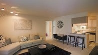 This single-floor space presents a thoughtfully designed open-concept layout that seamlessly connects the living area to the kitchen. The living room is anchored by a plush, light-toned sofa adorned with decorative cushions, complemented by a sleek dark coffee table resting atop a rich, dark area rug. A statement sunburst mirror serves as a striking focal point on the clean white walls, adding a touch of elegance alongside coordinating wall art. A compact accent bench provides additional seating near the hallway entrance, enhancing the room's functionality. The kitchen area features a raised bar-height counter with dark barstools, creating a casual dining and entertaining space. Neutral-toned cabinetry and appliances are visible beyond the counter, suggesting a well-equipped culinary space. Light-toned carpeting flows throughout the living area, contributing to the warm and inviting ambiance. A hallway visible in the background leads to additional rooms, hinting at a practical and efficient floor plan. The overall aesthetic is modern and cohesive, with a carefully curated mix of dark and light furnishings set against a soft, neutral backdrop.