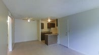 This single-floor unit offers a practical and functional open-concept layout that seamlessly connects the living area to the kitchen. The living space is generously proportioned, finished with light-toned carpeting and a ceiling fan that enhances air circulation and comfort. A textured popcorn ceiling runs throughout, lending a classic, familiar character to the interior.

The kitchen is efficiently designed and positioned at the heart of the unit, featuring dark wood cabinetry that provides ample storage space. A kitchen peninsula serves as a natural divider between the cooking and living areas while offering additional counter space and functionality. The kitchen is equipped with a white refrigerator and includes an under-cabinet range hood, with a sink positioned along the back wall beneath the upper cabinets.

The entry door is conveniently located adjacent to the kitchen, providing direct access to the main living area. The overall color palette is neutral and understated, with white walls and light flooring creating a bright, clean atmosphere throughout. The layout is straightforward and well-organized, making efficient use of the available space while maintaining an open and airy feel ideal for everyday living.