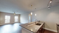 Located at 2701 Old Mayport Rd in Jacksonville, FL, this single-floor residence offers a well-designed open-concept layout that seamlessly connects the kitchen and living areas. The kitchen is a standout feature, showcasing rich dark cabinetry, granite countertops with a mosaic tile backsplash, stainless steel appliances including a refrigerator, microwave, and range, and a large peninsula with an undermount sink and elegant pendant lighting overhead. Track lighting further enhances the kitchen's functionality and style. The spacious living area is finished with dark hardwood-style flooring, neutral-toned walls, and a ceiling fan, with natural light streaming in through multiple windows fitted with blinds. The bedroom features plush carpeting, a ceiling fan, and direct access to a private balcony through a glass-paneled door. The bathroom is finished with a dark vanity, white countertop, large mirror, and a tiled tub-shower combination with modern chrome fixtures. A dedicated in-unit laundry closet houses a full-size washer and dryer alongside a water heater, adding everyday convenience. Throughout the space, clean lines, consistent neutral tones, and quality finishes create a cohesive and comfortable living environment.