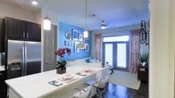 This single-floor space showcases a thoughtfully designed open-concept layout that effortlessly blends the dining and living areas into one cohesive environment. A striking blue accent wall serves as the visual centerpiece of the living area, complemented by a curated gallery wall of framed artwork and a decorative mirror that adds depth and personality to the space.

The dining area features a sleek white bar-style counter with wire accent chairs, illuminated by modern pendant lights that hang at varying heights, creating an inviting atmosphere for casual dining and entertaining. Dark hardwood flooring runs throughout, providing a rich contrast to the bright, airy color palette.

The living area is anchored by a light-toned sofa adorned with colorful throw pillows, positioned beneath a ceiling fan that ensures year-round comfort. Patterned drapes frame large windows that allow generous natural light to pour in, highlighting the open and welcoming ambiance of the room.

Subtle design details — including fresh floral arrangements, layered area rugs, and carefully chosen décor — lend the space a polished yet approachable character. The overall aesthetic balances bold color choices with clean, modern furnishings, resulting in a dynamic and functional living environment.