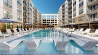 Located at 2000 Aureum Drive in Franklin, Tennessee, this exceptional outdoor amenity space is set within a contemporary multi-story residential community defined by a striking blend of warm brick and modern white façades. At the heart of the space is a generously sized resort-style swimming pool with crystal-clear blue water, outfitted with in-pool lounge chairs and complemented by rows of white chaise lounges and navy-striped umbrellas along the sundeck. A shallow tanning shelf offers additional in-water relaxation options. Surrounding the pool, the courtyard is thoughtfully designed with multiple distinct zones. A dedicated outdoor lounge area features stylish woven seating arranged around a central fire pit, offering a comfortable social setting. An outdoor kitchen and bar area — complete with grills, a prep counter, and bar seating — provides an ideal space for entertaining. A pergola-shaded retreat adds a sense of intimacy and shade. Paved pathways wind through curated landscaping with ornamental trees, trimmed shrubs, and stone accents. A large outdoor screen serves as a focal point for communal gathering. The overall ambiance is polished and resort-inspired, seamlessly merging indoor-outdoor living within the vibrant Franklin, TN community.