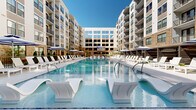 Located at 2000 Aureum Drive in Franklin, Tennessee, this exceptional outdoor amenity space is set within a contemporary multi-story residential community defined by a striking blend of warm brick and modern white façades. At the heart of the space is a generously sized resort-style swimming pool with crystal-clear blue water, outfitted with in-pool lounge chairs and complemented by rows of white chaise lounges and navy-striped umbrellas along the sundeck. A shallow tanning shelf offers additional in-water relaxation options. Surrounding the pool, the courtyard is thoughtfully designed with multiple distinct zones. A dedicated outdoor lounge area features stylish woven seating arranged around a central fire pit, offering a comfortable social setting. An outdoor kitchen and bar area — complete with grills, a prep counter, and bar seating — provides an ideal space for entertaining. A pergola-shaded retreat adds a sense of intimacy and shade. Paved pathways wind through curated landscaping with ornamental trees, trimmed shrubs, and stone accents. A large outdoor screen serves as a focal point for communal gathering. The overall ambiance is polished and resort-inspired, seamlessly merging indoor-outdoor living within the vibrant Franklin, TN community.