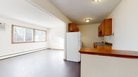 This single-floor residential unit offers a practical and functional layout with a clean, neutral aesthetic throughout. The open-concept design connects the kitchen directly to the living area, creating a cohesive and versatile shared space. The kitchen is equipped with warm-toned wooden cabinetry, a granite-style countertop, a double sink, and a full-size refrigerator. A peninsula counter separates the kitchen from the adjoining living space, which features durable hardwood-style flooring and a wall-mounted air conditioning unit alongside baseboard heating for year-round climate control. Natural light flows generously into the living area through wide windows fitted with horizontal blinds, offering views of the surrounding neighborhood. The bedroom is carpeted for added comfort and includes a spacious closet with built-in shelving, as well as a bright window that allows ample daylight to fill the room. The full bathroom is finished with classic white square wall tiles accented by blue tile details, complemented by a mosaic tile floor, a soaking tub, and a mirrored vanity cabinet. Consistent warm wood trim and baseboards tie the interior together, giving the unit a cohesive and well-maintained appearance throughout.