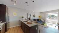 This single-floor residence is designed with a contemporary aesthetic, combining functional layouts with modern finishes. The open-concept kitchen and living area serves as the heart of the space, anchored by a large granite-topped island with an undermount sink and seating for multiple guests. Pendant lighting hangs above the island, adding a stylish focal point. The kitchen is outfitted with dark espresso cabinetry, a tile backsplash, and a full suite of stainless steel appliances, including a range, microwave, and refrigerator. Light hardwood-style flooring runs throughout, lending warmth and continuity across all areas.

The living area flows naturally from the kitchen and features a sliding glass door that allows natural light to fill the space and provides access to the outdoors. Two bedrooms offer comfortable retreats; one features a dedicated workspace with a leaning shelf unit, while the primary bedroom includes direct access to an en-suite bathroom equipped with a glass-enclosed walk-in shower. A second full bathroom features a soaking tub with full tile surround. Neutral gray walls throughout create a clean, cohesive backdrop well-suited to a variety of interior styles.