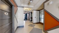 This thoughtfully designed single-floor amenity space offers a seamless blend of functionality and contemporary style. The entry corridor sets the tone with its striking geometric décor — bold orange accent panels, gray trim detailing, and a large-format checkerboard tile floor — leading guests into the heart of the space.

The expansive open-plan lounge and dining area is a standout feature, characterized by tray ceilings with dark trim, large floor-to-ceiling windows that invite abundant natural light and outdoor views, and light-toned large-format tile flooring. A sputnik-style chandelier adds an elegant focal point overhead. This versatile gathering space accommodates multiple seating configurations, from casual lounge sofas to formal dining tables with modern chairs.

A dedicated media room offers a more intimate setting, furnished with patterned swivel chairs, accent side tables, and a wall-mounted flat-screen TV, ideal for relaxed viewing.

The kitchenette area features rich dark wood cabinetry, a stainless steel sink, under-counter appliances, and glass display shelving set against a white brick accent wall with a contrasting dark ceiling — adding a refined, upscale feel.

A business center provides multiple computer workstations set within a bright, organized room accented with warm orange wall panels and ample natural light from a central window.