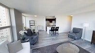 This single-floor space showcases a thoughtfully designed open-concept layout that seamlessly connects the living, dining, and kitchen areas into one cohesive and functional environment. Light-toned hardwood flooring runs throughout, lending the space a clean, contemporary feel and reflecting natural light that streams in through large windows along the perimeter.

The living area is anchored by a pair of plush charcoal armchairs arranged around a glass-topped coffee table, creating an inviting and well-appointed lounge space. A neutral color palette of whites and greys lends a sense of calm and sophistication throughout. Adjacent to the living space, a compact dining area with a round table and chairs offers a cozy spot for meals.

The kitchen is a standout feature, finished with dark espresso cabinetry, stainless steel appliances, and a white countertop island complete with seating for two. Track lighting above the kitchen provides focused illumination, while a textured white ceiling adds subtle visual interest throughout the space.

The overall ambiance is modern and airy, with a neutral, move-in-ready aesthetic that balances comfort and style. The open sightlines from the living area through to the kitchen and dining space make the unit feel expansive and well-connected.