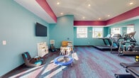 This fitness center in Shakopee, MN offers a well-equipped, single-floor exercise space designed to serve a range of wellness needs. The interior is immediately distinguished by its bold color palette — vivid teal walls accented by striking magenta banding along the upper walls and ceiling — creating an energetic and motivating atmosphere. Colorful patterned carpet in complementary blue and purple tones covers the floor throughout, adding to the lively aesthetic.

The space is generously outfitted with a variety of cardio machines, including treadmills, ellipticals, and stationary bikes, positioned to take advantage of the multiple windows that allow abundant natural light and views of the surrounding neighborhood. A dedicated strength training area features a multi-function cable machine and a well-stocked dumbbell rack, providing options for a full-body workout.

A vaulted ceiling with recessed lighting adds a sense of openness and height to the room. A wall-mounted flat-screen television and a water fountain are also present, enhancing convenience and comfort during workouts.

Notably, a children's play zone is integrated into the space, complete with a mini trampoline, ride-on toys, a play kitchen, and a small table and chairs — making this fitness center accessible and family-friendly. Large glass entry doors further brighten the space and provide easy access.