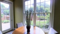 This single-story space includes a dining area that serves as a highlight of the interior. A large corner window arrangement, framed in crisp white multi-pane casings, floods the space with natural light and offers an unobstructed view of a well-maintained outdoor garden. The garden beyond features lush ground cover, neatly trimmed hedges, and mature trees, creating a serene green backdrop visible from within. The warm olive-toned walls complement the natural wood tones of the round dining table, which is centered beneath the windows and styled with a tall decorative arrangement of sculptural reed stems in a dark vase. The upholstered dining chair visible in the frame adds a clean, understated elegance. The overall ambiance of the space is calm and refined, blending classic architectural details—such as the traditional window grid pattern—with a comfortable, livable aesthetic. The abundance of natural light and direct visual connection to the outdoors gives the dining area an open, airy quality that enhances the sense of space throughout.