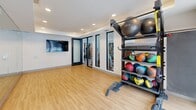 Located at 5505 W Northgate Rd in Rogers, Arkansas, this contemporary fitness center is thoughtfully organized across three distinct exercise rooms, offering a comprehensive workout environment for all fitness levels. The main gym area commands attention with soaring double-height ceilings accented by a striking geometric grid ceiling feature with integrated lighting. Large windows line the walls, flooding the space with natural light and providing views toward an outdoor pool area. The patterned carpet flooring and an extensive array of equipment — including cable machines, power racks, kettlebells, barbells, and benches — make this a well-equipped strength training hub. A dedicated cardio room features treadmills and elliptical machines beneath high ceilings with additional window views. A separate functional training studio showcases hardwood-style flooring, full-length mirrored walls, recessed lighting, wall-mounted TVs, and a curated selection of medicine balls, stability balls, and resistance tools. Glass partition walls and black-framed doors connect the rooms, maintaining an open, airy feel throughout while providing visual separation between zones. The overall design is clean, modern, and purposefully laid out to support a full range of fitness activities.