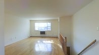 This single-floor space presents a clean, open layout centered around a well-proportioned main living area. The room is finished with gleaming hardwood floors that reflect natural light streaming in through a prominent double window along the far wall, creating a bright and airy atmosphere throughout. The walls are painted in a neutral, creamy white tone, lending the space a fresh and versatile backdrop suitable for a variety of interior styles.

A wall-mounted air conditioning unit sits beneath the window, complemented by a baseboard heating system that runs along the perimeter of the room, ensuring year-round climate comfort. The ceiling height is standard, contributing to a comfortable and functional sense of scale within the living area.

A classic wood-railed staircase with a turned newel post is positioned along one side of the room, adding a traditional architectural detail and serving as a notable design element. A doorway or passage is visible along the adjacent wall, suggesting additional rooms or spaces beyond the main living area. Overall, the space combines practical features with timeless finishes, offering a bright and welcoming environment.