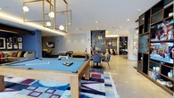 This single-floor community room is a thoughtfully designed, multi-functional gathering space that seamlessly blends recreation, socializing, and relaxation. The open layout is anchored by a prominent pool table set atop a bold, geometric area rug, complemented by a foosball table nearby, making the games area a central hub of activity. Geometric gold pendant lighting hangs overhead, adding a modern, upscale touch to the space.

Adjacent to the games zone, a casual dining and lounge area features sleek blue chairs gathered around round tables, ideal for conversations or casual meetings. A striking built-in shelving unit with deep navy blue tones serves as both a functional and decorative focal point, displaying curated décor and books. The open shelving divides the space while maintaining a sense of flow and connectivity throughout.

Further zones offer additional seating arrangements, including plush lounge furniture in warm yellow and gray tones, providing comfortable spots for residents to unwind. A dedicated children's play area with colorful furnishings adds inclusive versatility to the room. Throughout, the design palette of blues, golds, and neutral tones creates a cohesive, contemporary ambiance. Recessed lighting and polished concrete-style flooring enhance the modern aesthetic of this well-appointed community space.