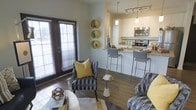 This single-floor space showcases an open-concept layout that seamlessly connects the living area and kitchen, creating a cohesive and functional environment. The living area is anchored by hardwood-style flooring and features patterned accent chairs with bold yellow throw pillows, adding a pop of color against neutral walls. Three decorative round wall shelves in a golden finish serve as a distinctive design element. French doors with horizontal blinds line one wall, allowing generous natural light to fill the space and providing access to the outdoors. The kitchen is well-appointed with white cabinetry, granite-style countertops, stainless steel appliances including a microwave and range, and a breakfast bar with modern bar stools. Pendant lighting and a track light fixture illuminate the kitchen, complementing the overall brightness of the space. The open flow between the living and kitchen areas makes the layout feel expansive and ideal for both everyday living and entertaining. The cohesive color palette of whites, grays, and golden yellows throughout ties the space together with a contemporary and inviting aesthetic.