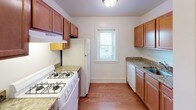 This single-floor unit offers a thoughtfully laid-out interior with warm hardwood flooring that runs consistently throughout the living spaces, lending a cohesive and inviting feel. The galley-style kitchen is a functional highlight, featuring rich wood cabinetry, granite-look countertops, a gas range, a full-size refrigerator, and a dishwasher. A window above the sink area brings in natural light, making the kitchen feel open and practical for everyday use. The spacious living area is bathed in natural light through large multi-pane windows and is equipped with a ceiling fan for year-round comfort. A distinctive set of dark wooden bi-fold doors adds a touch of character to the space. The bedrooms are generously sized, each featuring hardwood floors, ceiling fans, and ample natural light from well-placed windows. One bedroom includes a built-in closet with hanging rod storage. Classic cast-iron radiators found in several rooms speak to the building's architectural heritage while providing efficient heating. The bathroom is cleanly finished with tile surround, a shower-tub combo, a granite-topped vanity, and a mirrored medicine cabinet. Overall, the unit balances classic charm with functional, updated finishes.