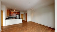 This single-floor residence features a thoughtfully designed open-concept layout that seamlessly connects the living and kitchen areas. Rich hardwood flooring flows throughout the main living space, creating a warm and cohesive aesthetic. The kitchen is well-appointed with warm-toned wood cabinetry, dark granite countertops, and a full suite of stainless steel appliances, including a gas range, built-in microwave, dishwasher, and refrigerator. A breakfast bar provides additional workspace and casual seating. Track lighting illuminates both the kitchen and living area, complementing the recessed ceiling lights throughout.

The living area is bright and open, leading to a sliding glass door that opens to a private balcony with city skyline views. The bedroom features plush carpeting and expansive floor-to-ceiling windows that frame impressive urban views, filling the room with natural light. The bathroom offers a generous vanity with granite countertops, warm wood cabinetry, a large mirror, and tile flooring, providing a clean and functional finish. Overall, the space conveys a modern, urban aesthetic with quality finishes throughout.