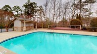 Located at 3374 Starboard Way in Fayetteville, NC, this community amenity facility offers a generously sized outdoor pool area encompassing over 3,300 square feet of usable space. The centerpiece is a large, geometric swimming pool with a classic blue tile border, entry steps with handrails, and clearly marked depth indicators, designed to accommodate a wide range of swimmers.

The pool is flanked by a spacious concrete deck that provides ample room for lounge seating, with multiple patio chairs positioned along the pool's edge for relaxation. A charming pool house anchors one side of the facility, featuring craftsman-inspired architecture with stone column bases, a covered outdoor pavilion with ceiling-mounted lighting, large multi-pane windows, and a dark pitched roof. The pavilion provides shaded gathering space directly adjacent to the pool.

On the opposite end of the deck, an elevated wooden platform area offers additional recreational space and overlooks the surrounding landscape. The entire facility is enclosed by a black wrought iron fence, providing both security and clear sightlines to the surrounding mature pine and hardwood trees that create a natural, serene backdrop. The lush wooded perimeter enhances privacy and lends a peaceful, retreat-like atmosphere to this well-appointed community space.