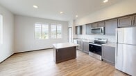 This single-floor unit on Centennial Trail in Arlington, Washington, offers a thoughtfully designed layout that maximizes its compact footprint. Light-toned luxury vinyl plank flooring runs throughout, lending a clean, cohesive aesthetic to the entire space.

The kitchen serves as a central highlight, featuring dark espresso-toned cabinetry with modern hardware, a full suite of stainless steel appliances including an electric range, microwave, dishwasher, and refrigerator, and a tile backsplash accent. A central island with a white quartz-style countertop provides additional workspace and casual seating potential. Recessed lighting illuminates the kitchen efficiently.

The living area is bathed in natural light through a trio of large windows that frame views of the surrounding Arlington landscape, including glimpses of distant mountains. Neutral white walls and clean trim throughout reinforce a bright, open atmosphere.

The bathroom doubles as a dedicated laundry room, housing a stacked washer and dryer unit alongside standard bathroom fixtures, making practical use of available space. Throughout the unit, white interior doors, recessed lighting, and consistent finishes contribute to a polished, move-in-ready presentation.
