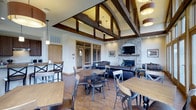 This single-floor space blends rustic and contemporary design elements to create a warm, inviting communal environment. The interior is anchored by dramatic vaulted ceilings with exposed dark wood beam detailing, which draw the eye upward and contribute to the spacious, airy atmosphere. Cylindrical pendant and drum lighting fixtures complement the architectural woodwork, casting a warm glow throughout.

The main gathering area is organized into distinct zones. A lounge area features dark leather sofas arranged around a stacked-stone fireplace with a wood mantle, providing a cozy focal point with a wall-mounted television above. Adjacent to this, multiple high-top dining tables with cross-back chairs offer casual seating, while a bar-height counter along one wall adds additional functionality. Wood-look flooring runs throughout, tying the zones together cohesively.

Large windows and glass doors connect the interior to an expansive outdoor terrace, where bistro-style high-top tables and chairs are arranged for al fresco seating. The terrace offers open views of surrounding greenery and neighboring buildings. The exterior façade features a combination of warm wood-tone siding and large arched windows, reinforcing the lodge-inspired aesthetic of the overall space.