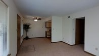 This single-floor residence offers a practical and comfortable layout designed for everyday living. The main living area is spacious and open, finished with neutral wall-to-wall carpeting that adds warmth underfoot. A ceiling fan with a light fixture is centrally positioned, providing both air circulation and ambient lighting throughout the space. Dark wood baseboards provide a grounding contrast to the light-colored walls, lending the interior a clean, classic aesthetic.

The living area flows naturally toward a partially open kitchen, visible through a connecting passage. The kitchen features wood-toned upper cabinetry and a built-in drawer unit below, offering ample storage for everyday essentials. The warm cabinetry tones complement the neutral palette of the surrounding spaces.

Natural light enters through a window in the living area, where a decorative potted tree adds a touch of greenery and life to the room. A ventilation register is visible on the ceiling, indicating a central HVAC system for year-round climate control. A wooden interior door near the far wall provides access to an adjoining room or hallway, completing the single-level floor plan. Overall, the space presents a straightforward, well-maintained interior suited for comfortable day-to-day living.