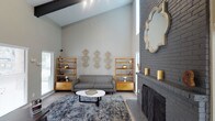 This single-story Dallas home blends contemporary design with warm, inviting character. The living area immediately captures attention with its dramatic vaulted ceilings accented by exposed dark beams, creating an impressive sense of vertical space and openness. Recessed track lighting highlights the room's architectural lines, while multiple windows allow natural light to pour in from multiple angles.

A standout feature of the living space is the floor-to-ceiling painted brick fireplace, finished in a sophisticated dark gray that adds texture and a bold focal point to the room. Flanking a sleek gray sofa, a pair of warm-toned wooden shelving units provide both storage and display space, complemented by curated gold accents and decorative wall art. A round white coffee table anchors the seating area atop a richly patterned area rug laid over light hardwood-style flooring.

The overall ambiance is modern yet cozy — a thoughtful balance of clean lines, natural materials, and warm metallic touches. The neutral gray and white color palette throughout the living space lends a calm, sophisticated feel, while the architectural details and carefully selected furnishings give the home a distinct, curated character well-suited to the Dallas aesthetic.
