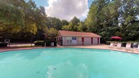 This single-story community amenity space offers residents two primary facilities: a generously sized outdoor swimming pool and a fully equipped fitness center. The outdoor pool area is a standout feature, showcasing a large, turquoise pool enclosed by a wrought-iron fence and framed by a lush canopy of mature trees that provide natural shade and a serene atmosphere. Poolside lounge chairs and umbrella-shaded seating areas offer comfortable spaces to relax. A dedicated pool house sits at one end of the pool, featuring distinct red-accented doors and housing restroom facilities for pool guests. The fitness center is housed within the same building complex and is thoughtfully designed with vaulted ceilings, recessed lighting, and a ceiling fan for comfort. The space is painted in a calming two-tone light blue palette and features large windows and French doors that invite abundant natural light and views of the surrounding greenery. The gym is well-appointed with multiple treadmills, elliptical trainers, stationary and recumbent bikes, a multi-function cable weight machine, and additional strength equipment. A floor-to-ceiling mirror wall enhances the sense of space, while a wall-mounted television adds convenience for cardio workouts.