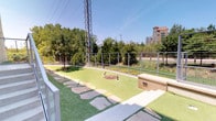 This single-level outdoor amenity space in Dallas is thoughtfully designed to offer a well-appointed recreational environment adjacent to a light brick building. The grounds are covered in vibrant artificial turf, providing a clean, low-maintenance surface throughout the area. A winding flagstone pathway meanders across the lawn, creating a natural flow through the space and connecting key areas with a rustic yet refined aesthetic.

A dedicated putting green occupies one section of the grounds, making this an ideal spot for casual outdoor recreation. Decorative rock beds and low-profile landscape lighting are strategically placed throughout, adding texture and visual interest. Raised concrete steps with sleek stainless steel and glass railings provide access between the building and the outdoor area, emphasizing a modern design sensibility.

The perimeter is enclosed by a contemporary cable-wire fence with metal posts, maintaining an open sightline to the surrounding urban greenery, mature trees, and city buildings beyond. A waste station is also present, indicating pet-friendly accommodations. The space receives abundant natural sunlight and offers an open-air ambiance that blends urban convenience with thoughtfully landscaped outdoor living.
