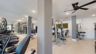 Located at 3701 Southwest 34th Street in Gainesville, Florida, this fitness center is a thoughtfully designed exercise facility that offers a comprehensive workout environment within a single, open floor. The space features light wood-tone flooring in the primary workout zone, transitioning to a polished tile surface near the entrance, and is kept bright and airy through a generous bank of floor-to-ceiling windows along one wall that invite abundant natural light and outdoor views.

The cardio section is equipped with multiple treadmills, elliptical trainers, and stationary bikes positioned to take advantage of the natural light and window views. A full-length mirrored wall amplifies the sense of openness while allowing users to monitor their form. Overhead, sleek ceiling fans maintain comfortable air circulation throughout the space.

The strength training area is well-appointed with a variety of cable and weight machines, adjustable benches, a dumbbell rack, and a dedicated functional fitness station stocked with medicine balls, resistance bands, and exercise balls on wall-mounted storage racks. Structural columns are integrated cleanly into the design, finished in a neutral gray tone that complements the modern aesthetic.

Decorative touches, including botanical moss wall art and colorful framed prints, add a polished, welcoming ambiance. A teal built-in storage credenza near the entrance provides a practical organizational element. The facility is finished with recessed lighting throughout, ensuring consistent illumination across all workout zones.