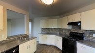 This single-floor space showcases a well-appointed kitchen as its central feature. The kitchen is designed with ample white cabinetry spanning both upper and lower sections, providing generous storage capacity. Dark laminate countertops offer a striking contrast against the white cabinet faces, while a mosaic tile backsplash in earthy tones of grey, brown, and beige adds visual texture and a polished finish to the cooking area.

The kitchen is equipped with a black range and overhead ventilation hood, complementing the overall dark accent theme. A double-basin sink is positioned along a secondary counter run, providing practical workspace. The L-shaped counter layout maximizes usable prep and cooking area efficiently.

Dark hardwood-style flooring runs throughout the visible space, contributing a warm, cohesive feel underfoot. The neutral grey walls keep the atmosphere clean and understated, allowing the design elements to stand out. A hallway visible in the background suggests additional rooms beyond the kitchen, and flush-mount ceiling lighting ensures the space remains well-lit. Overall, the space presents a functional and contemporary aesthetic suitable for everyday living.